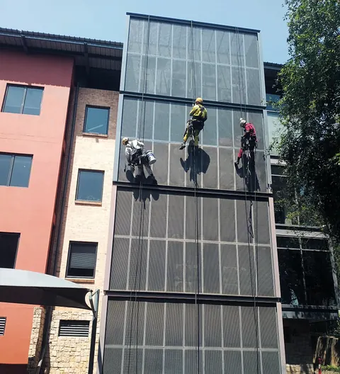 Three MP rope access technicians painting a perforated steel facade under a blue sky